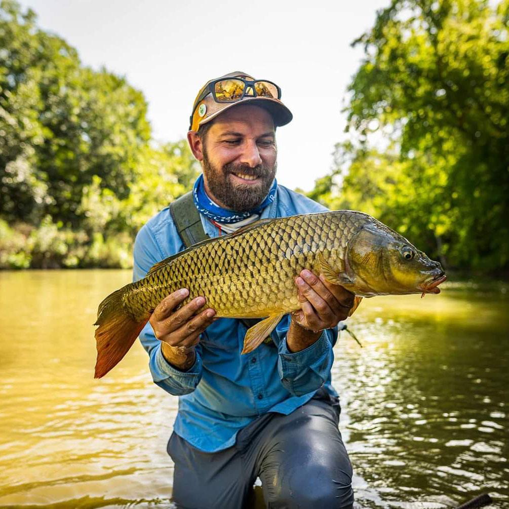 Lead guide Cory holding a common carp while kneeling in a river.